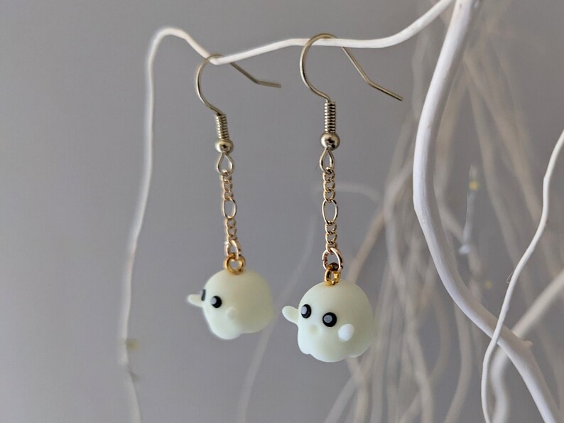 Op de afbeelding: Een paar oorbelletjes met een witte spookhanger met zwarte ogen. De spookhanger is bevestigd aan een gouden ketting en een zilveren haak.