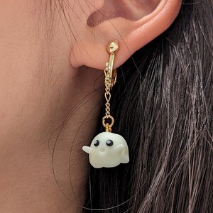 Op de afbeelding: Een paar gouden clip-on oorbellen met een witte spookhanger. Het spook heeft zwarte ogen en een kleine glimlach.