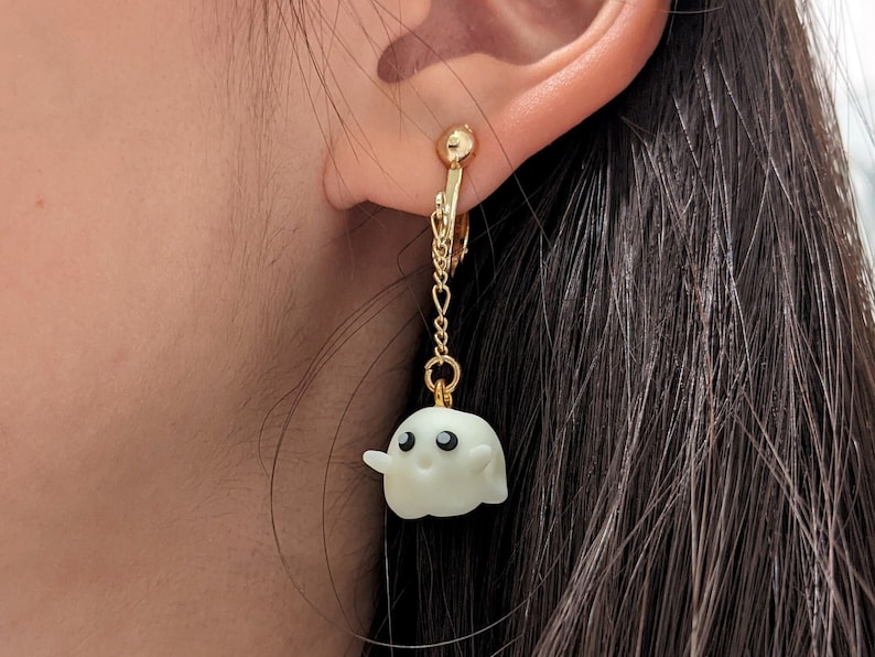 Op de afbeelding: Een paar gouden clip-on oorbellen met een witte spookhanger. Het spook heeft zwarte ogen en een kleine glimlach.