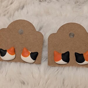 Op de afbeelding: Twee paar oorknopjes in de vorm van kattenkoppen, elk met een lapjeskatpatroon met zwarte, oranje en witte vlekken. De oorbellen zijn bevestigd op bruine, rechthoekige kartonnen displaykaarten met afgeronde bovenkanten en een rond gat.