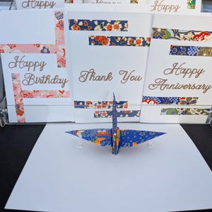 Op de afbeelding: Drie witte wenskaarten met bloemmotief washi-tape accenten en gouden cursieve tekst met Happy Birthday, Thank You en Happy Anniversary. Een blauwe origami kraanvogel met een kleurrijk bloemmotief staat voor de kaarten.
