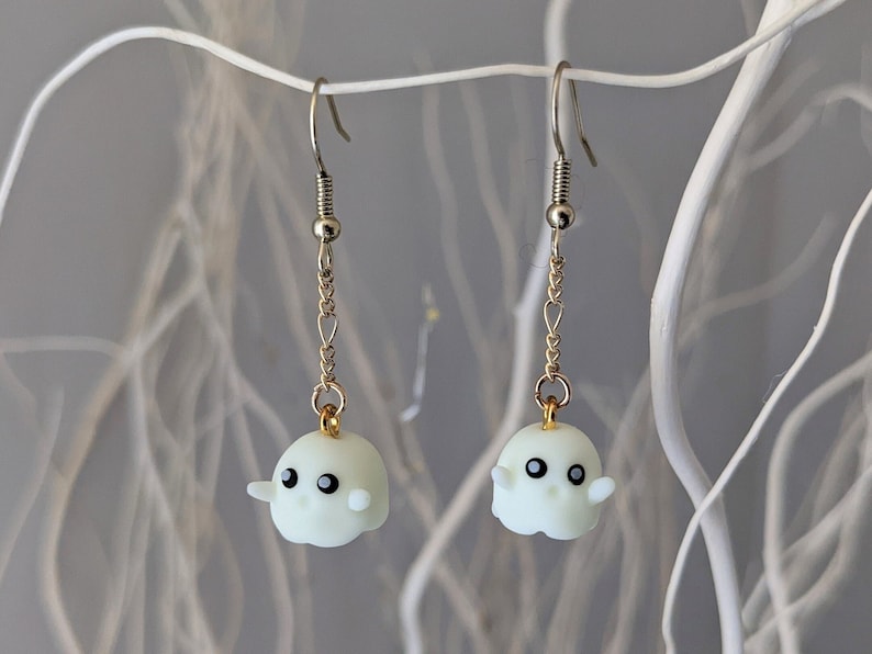 Op de afbeelding: Een paar zilveren en gouden oorbellen met witte spookbedels. De spoken hebben zwarte ogen en een kleine glimlach.