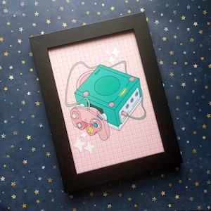 May include: A framed print of a pink and white grid pattern with a green and black video game console and a pink and black controller. The console has a green screen and the controller has a black button.