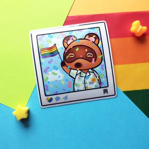 May include: A sticker featuring a cartoon raccoon holding a rainbow flag in front of a blue background with confetti. The sticker is in the shape of a social media post with a heart, comment bubble, and paper airplane icon.