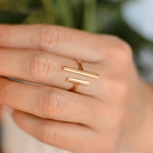 Op de afbeelding: Een gouden ring met een minimalistisch ontwerp met twee parallelle staven. De ring wordt aan een vinger gedragen.