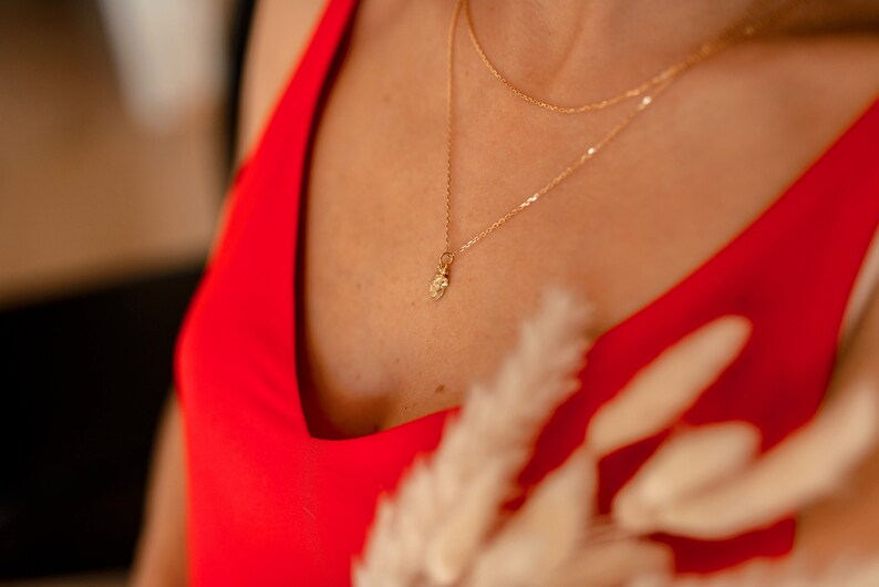 May include: A close-up of a person wearing a red v-neck top with a gold chain necklace featuring a small pendant. The necklace is layered with a second, thinner chain.