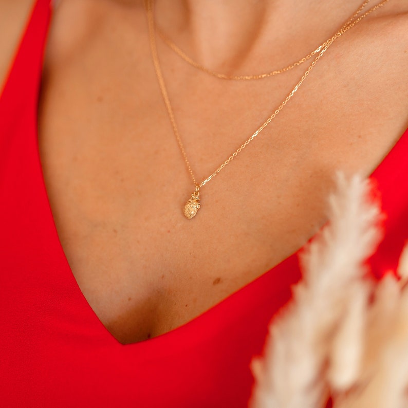 May include: A gold necklace with a small pineapple pendant. The necklace is layered on a person wearing a red top.