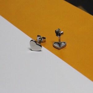 以下が含まれることがあります： 銀色のハート型のスタッドピアスのペア。ピアスは白と黄色の背景の上にあります。
