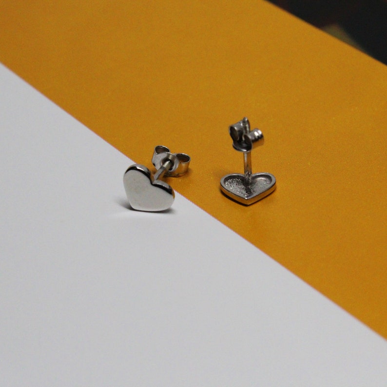 以下が含まれることがあります： 銀色のハート型のスタッドピアスのペア。ピアスは白と黄色の背景の上にあります。