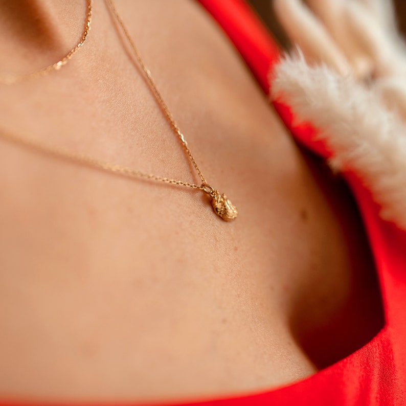 May include: A gold chain necklace with a small, textured, gold pendant. The necklace is worn by a person wearing a red top.