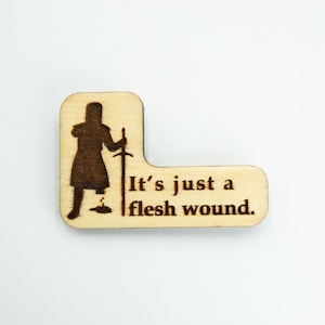May include: Wooden laser-cut pin featuring a silhouette of a knight with a missing leg, holding a sword. The text "It's just a flesh wound" is below the knight. The pin is light brown and has a unique shape.