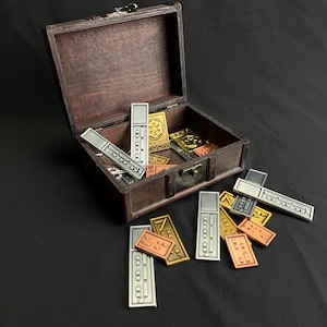 Könnte beinhalten: Eine dunkle Holzkiste voller Metallmarken in Silber-, Gold- und Kupfertönen. Die Marken sind rechteckig, quadratisch und dreieckig und weisen geätzte Muster auf. Einige Marken liegen außerhalb der Kiste auf einer dunklen Oberfläche.