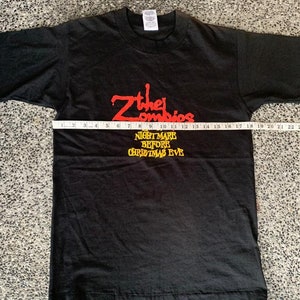 Peut inclure: T-shirt noir avec l'inscription rouge "Zther Zombies" et l'inscription jaune "NIGHTMARE BEFORE CHRISTMAS EVE". Un m&egrave;tre ruban est pos&eacute; sur le devant du t-shirt. Le t-shirt est &agrave; plat. Le t-shirt est de taille moyenne.