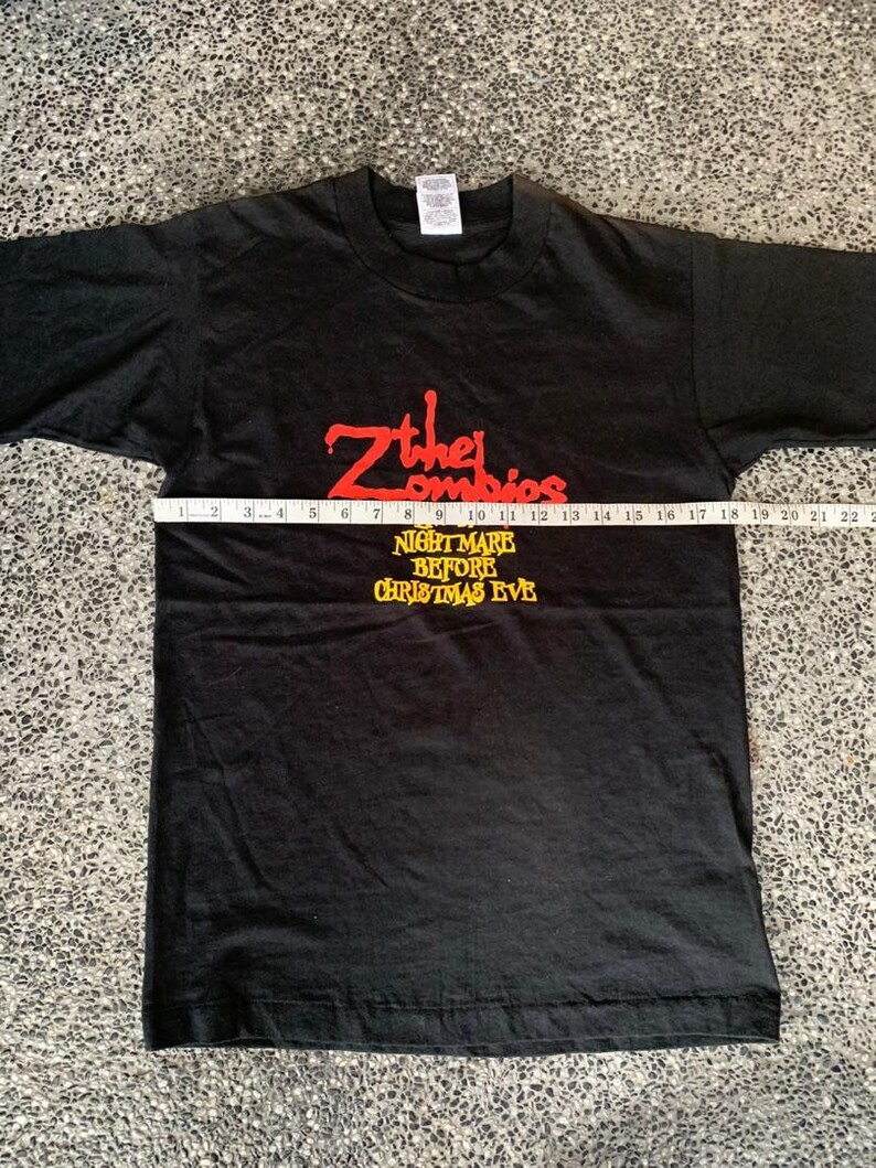 Peut inclure: T-shirt noir avec l'inscription rouge "Zther Zombies" et l'inscription jaune "NIGHTMARE BEFORE CHRISTMAS EVE". Un m&egrave;tre ruban est pos&eacute; sur le devant du t-shirt. Le t-shirt est &agrave; plat. Le t-shirt est de taille moyenne.