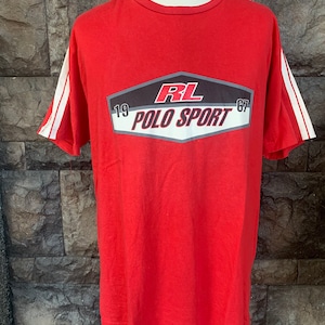 Op de afbeelding: Rood t-shirt met witte strepen op de mouwen. Het shirt heeft een afbeelding met de tekst "RL POLO SPORT 1967" in een zwart, wit en rood ontwerp. Het t-shirt wordt op een mannequin getoond.