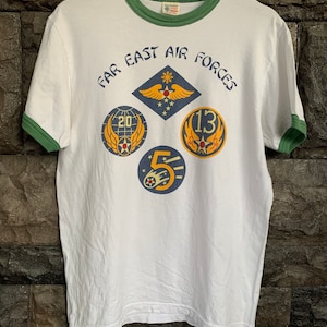 May include: White t-shirt with a green ringer and a graphic print of "Far East Air Forces" with four military insignia. The insignia are in blue, yellow, and red.