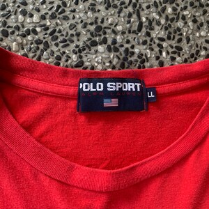 Op de afbeelding: Een rood Polo Sport T-shirt met een ronde hals. Het marineblauwe label heeft de tekst "POLO SPORT RALPH LAUREN" en een Amerikaanse vlag. Het shirt is maat LL.