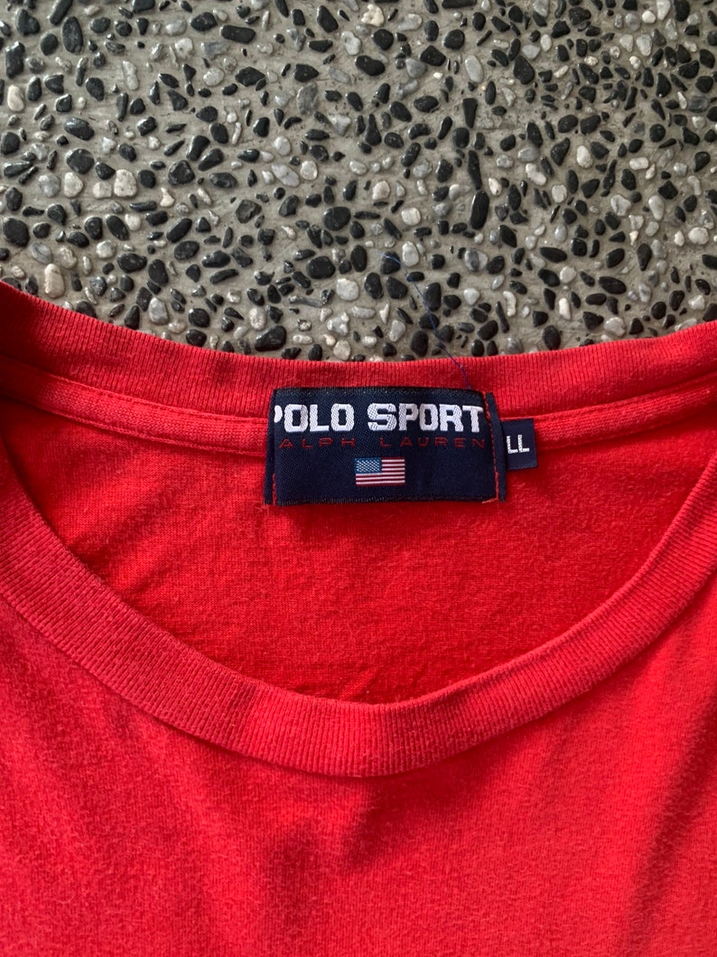 Op de afbeelding: Een rood Polo Sport T-shirt met een ronde hals. Het marineblauwe label heeft de tekst "POLO SPORT RALPH LAUREN" en een Amerikaanse vlag. Het shirt is maat LL.