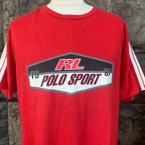 Op de afbeelding: Rood t-shirt met witte strepen op de mouwen. Het shirt heeft een afbeelding met de tekst "RL POLO SPORT 67" in wit en rood op een zwarte en grijze achtergrond.