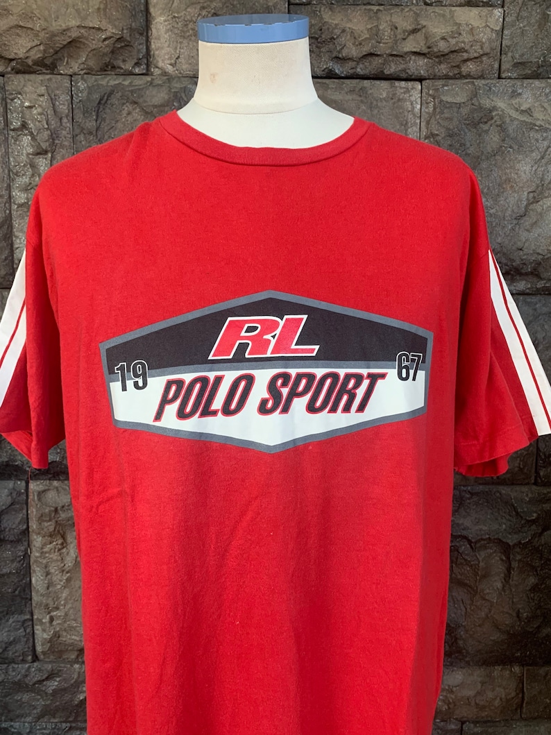 Op de afbeelding: Rood t-shirt met witte strepen op de mouwen. Het shirt heeft een afbeelding met de tekst "RL POLO SPORT 67" in wit en rood op een zwarte en grijze achtergrond.