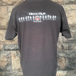 May include: Black t-shirt with the text "Black Flys WANTED" in white and red. The shirt features a graphic of silhouettes. The t-shirt is displayed on a mannequin.