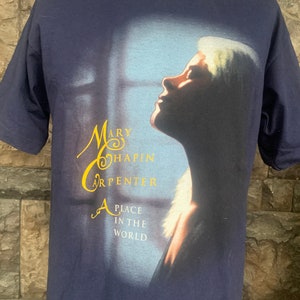 May include: A navy blue t-shirt featuring a faded image of Mary Chapin Carpenter. The image shows Carpenter looking upwards with her blonde hair blowing in the wind. The text "A Place in the World" is printed in gold beneath the image.