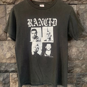 Puede incluir: Camiseta negra con una impresión blanca de la banda Rancid. La impresión presenta el nombre de la banda en una fuente gótica y cuatro fotos en blanco y negro de los miembros de la banda.
