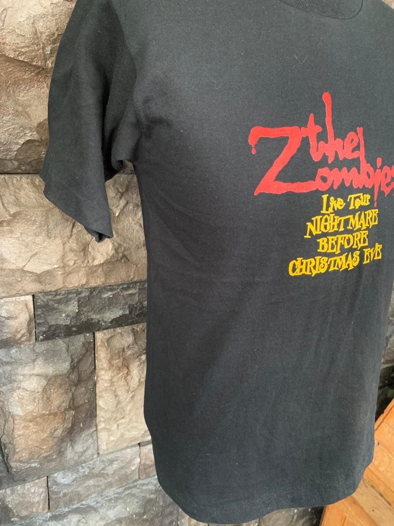 Peut inclure: T-shirt noir avec l'inscription "Z the Zombies" en rouge et "Live Tour! Nightmare Before Christmas Eve" en jaune. Le t-shirt est fait d'une mati&egrave;re douce et a des manches courtes.
