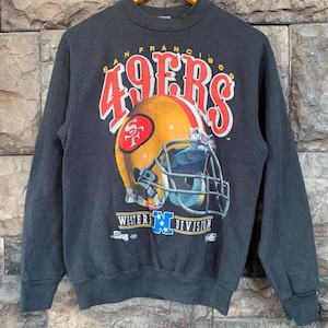 Könnte beinhalten: Ein schwarzes Langarm-Sweatshirt mit einer Grafik eines goldenen Football-Helms mit rotem Logo. Der Text lautet San Francisco 49ers Western Division mit einem blauen N-Logo. Das Design enthält rote und weiße Schriftzüge auf einem dunklen Stoff.