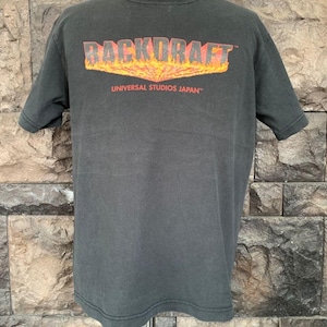 May include: Dark gray t-shirt featuring the text "BACKDRAFT" in orange and red flames above "UNIVERSAL STUDIOS JAPAN" in white. The short-sleeved shirt is displayed on a mannequin against a brick wall background.