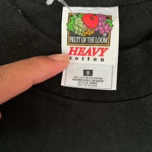 Peut inclure: Gros plan d'un t-shirt noir avec une &eacute;tiquette. L'&eacute;tiquette pr&eacute;sente le logo Fruit of the Loom avec une pomme rouge, des raisins et des feuilles. L'&eacute;tiquette indique "HEAVY cotton" et "S". Le t-shirt est en 100% coton.