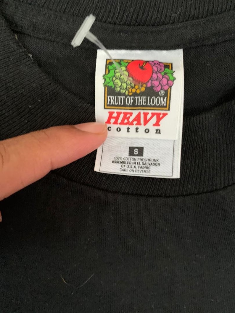 Peut inclure: Gros plan d'un t-shirt noir avec une &eacute;tiquette. L'&eacute;tiquette pr&eacute;sente le logo Fruit of the Loom avec une pomme rouge, des raisins et des feuilles. L'&eacute;tiquette indique "HEAVY cotton" et "S". Le t-shirt est en 100% coton.