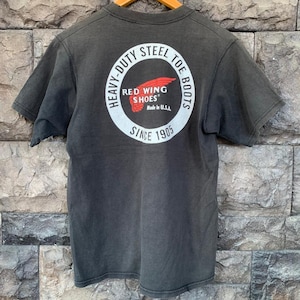 May include: Dark gray t-shirt with a circular graphic on the back. The graphic features the text "HEAVY-DUTY STEEL TOE BOOTS" around the perimeter, with "RED WING SHOES" and a red wing logo in the center. The text "Made in U.S.A." and "SINCE 1905" are also included.