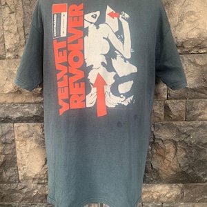 Könnte beinhalten: Ein dunkelgrünes T-Shirt mit einem weißen und roten Grafikdruck einer Frau und dem Text "VELVET REVOLVER" in Rot. Der Text "COPYRIGHTED" ist ebenfalls auf dem T-Shirt zu sehen.