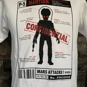 Puede incluir: Camiseta blanca con un gráfico negro y rojo de un marciano de la película "Mars Attacks!" El gráfico incluye un diagrama detallado de la anatomía y el equipo del marciano, incluyendo un arma de rayos, un traje espacial y un tubo de aire. El texto "CONFIDENTIAL" está impreso en letras rojas grandes en todo el gráfico. La parte inferior del gráfico incluye la fecha "12/20/96" y el número "29032028".