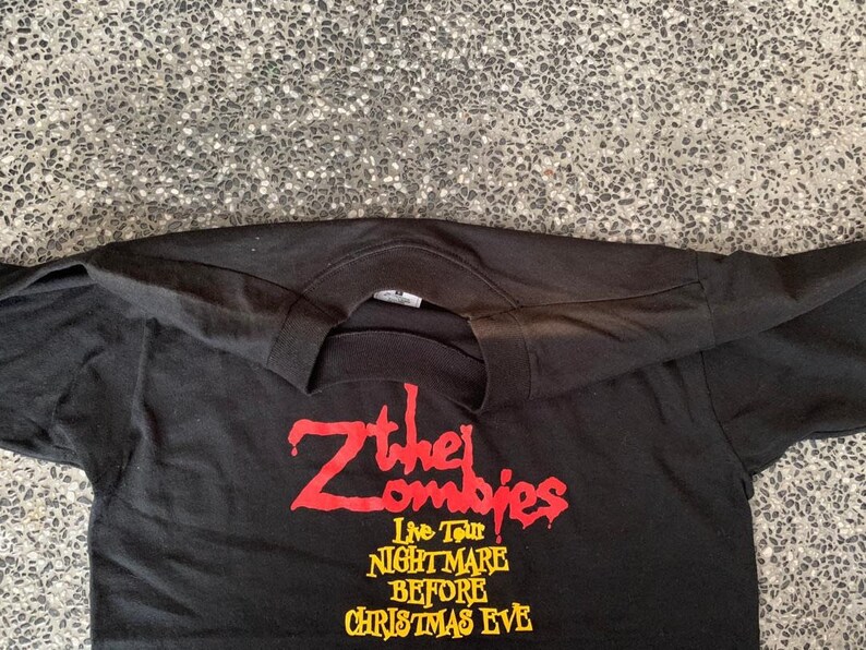 Peut inclure: T-shirt noir &agrave; manches longues avec l'inscription "Z the Zombies Live Tour Nightmare Before Christmas Eve" en rouge et jaune. Le t-shirt a un col rond et est fait d'une mati&egrave;re douce.