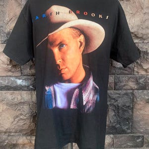 May include: A black short-sleeve t-shirt featuring a graphic print of a person wearing a cowboy hat and a plaid shirt. The text GARTH BROOKS is printed in a stylized font above the portrait. The shirt has a crew neckline and a straight hem.