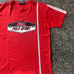 Op de afbeelding: Rood Polo Sport T-shirt met witte strepen op de mouwen. Het shirt heeft een afbeelding met de tekst "RL POLO SPORT 1967". Het shirt is ongeveer 76 cm lang.
