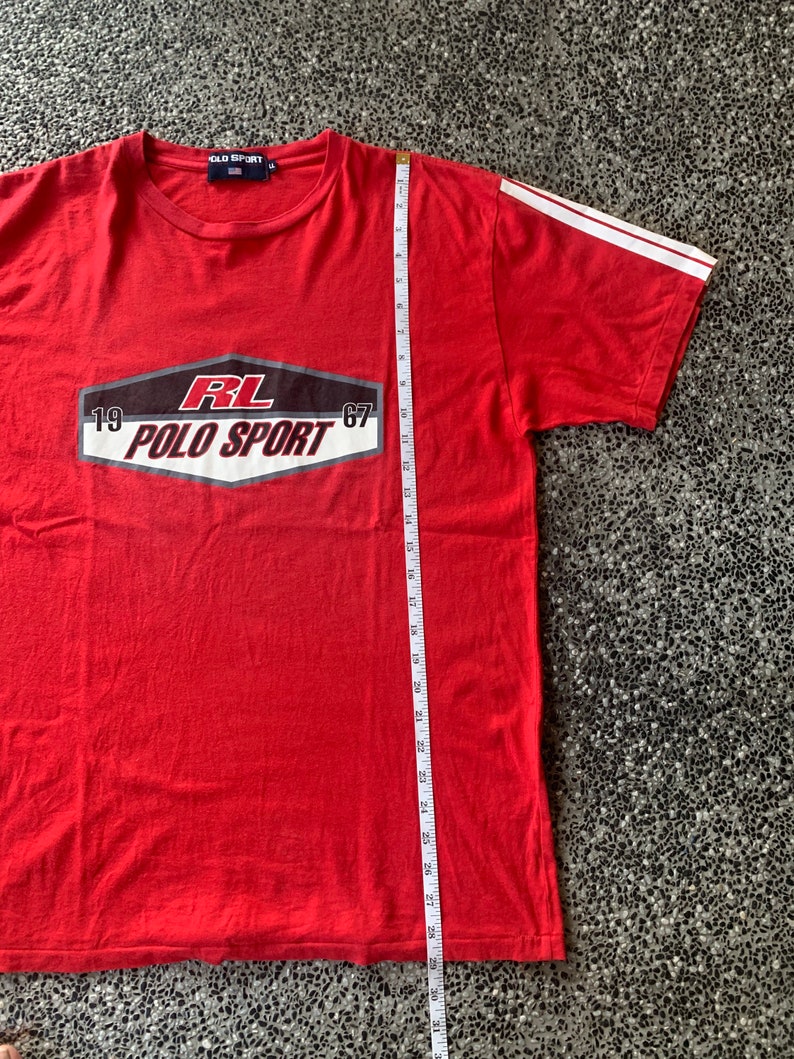 Op de afbeelding: Rood Polo Sport T-shirt met witte strepen op de mouwen. Het shirt heeft een afbeelding met de tekst "RL POLO SPORT 1967". Het shirt is ongeveer 76 cm lang.