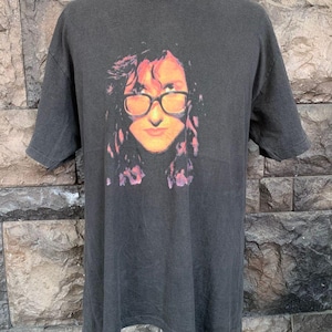 May include: Dark gray t-shirt featuring a graphic of a person wearing glasses. The graphic has orange, red, and purple tones. The t-shirt has short sleeves and a crew neck.