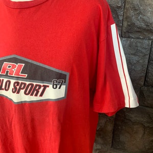 Op de afbeelding: Rood t-shirt met witte strepen op de mouw en een afbeelding op de voorkant. De afbeelding bevat de tekst "RL POLO SPORT 67" in wit, zwart en grijs.