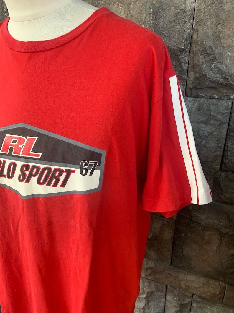 Op de afbeelding: Rood t-shirt met witte strepen op de mouw en een afbeelding op de voorkant. De afbeelding bevat de tekst "RL POLO SPORT 67" in wit, zwart en grijs.