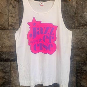 May include: White tank top with a pink and purple graphic that reads "Jazzercise Japan". The graphic is in a starburst shape. The tank top is hanging on a wooden hanger.