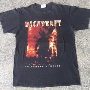 Peut inclure: T-shirt noir avec le titre du film "BACKDRAFT" en lettres rouges et blanches. Le motif comprend une image enflammée d'une personne. Le texte "UNIVERSAL STUDIOS" est imprimé en blanc en dessous.