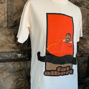以下が含まれることがあります： クリーム色のTシャツ。オレンジ色の長方形の中に、ファット・アルバートの漫画のイメージが描かれています。黒い縁取りがあります。「fat albert」の文字が茶色とオレンジ色でプリントされています。半袖で、クルーネックです。