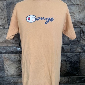 Puede incluir: Camiseta color beige con la palabra "Gonge" en escritura azul y un logo rojo, blanco y azul. La camiseta se exhibe en un maniquí. La camiseta de manga corta tiene cuello redondo. Fondo de pared de piedra texturizada.