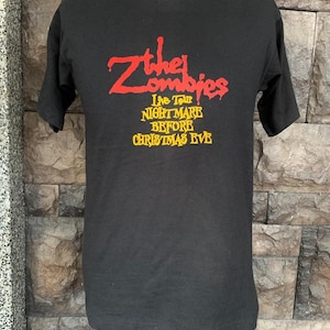 Peut inclure: T-shirt noir avec le nom du groupe "The Zombies" en rouge, et "Live Tour NIGHTMARE BEFORE CHRISTMAS EVE" en jaune. Le t-shirt est pr&eacute;sent&eacute; sur un mannequin devant un mur de briques.
