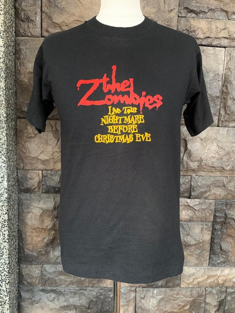 Peut inclure: T-shirt noir avec le nom du groupe "The Zombies" en rouge, et "Live Tour NIGHTMARE BEFORE CHRISTMAS EVE" en jaune. Le t-shirt est pr&eacute;sent&eacute; sur un mannequin devant un mur de briques.