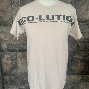 Op de afbeelding: Een beige T-shirt met een zwarte afbeelding met de tekst "ECO-LUTIO" en "LIKE MATCHES YOU PLAY WITH MATCHES, YOU SET"