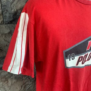 Op de afbeelding: Rood t-shirt met witte strepen op de mouw en een afbeelding op de voorkant. De afbeelding bevat het woord "POLO" en het nummer "19". Het shirt is gemaakt van een zacht materiaal.